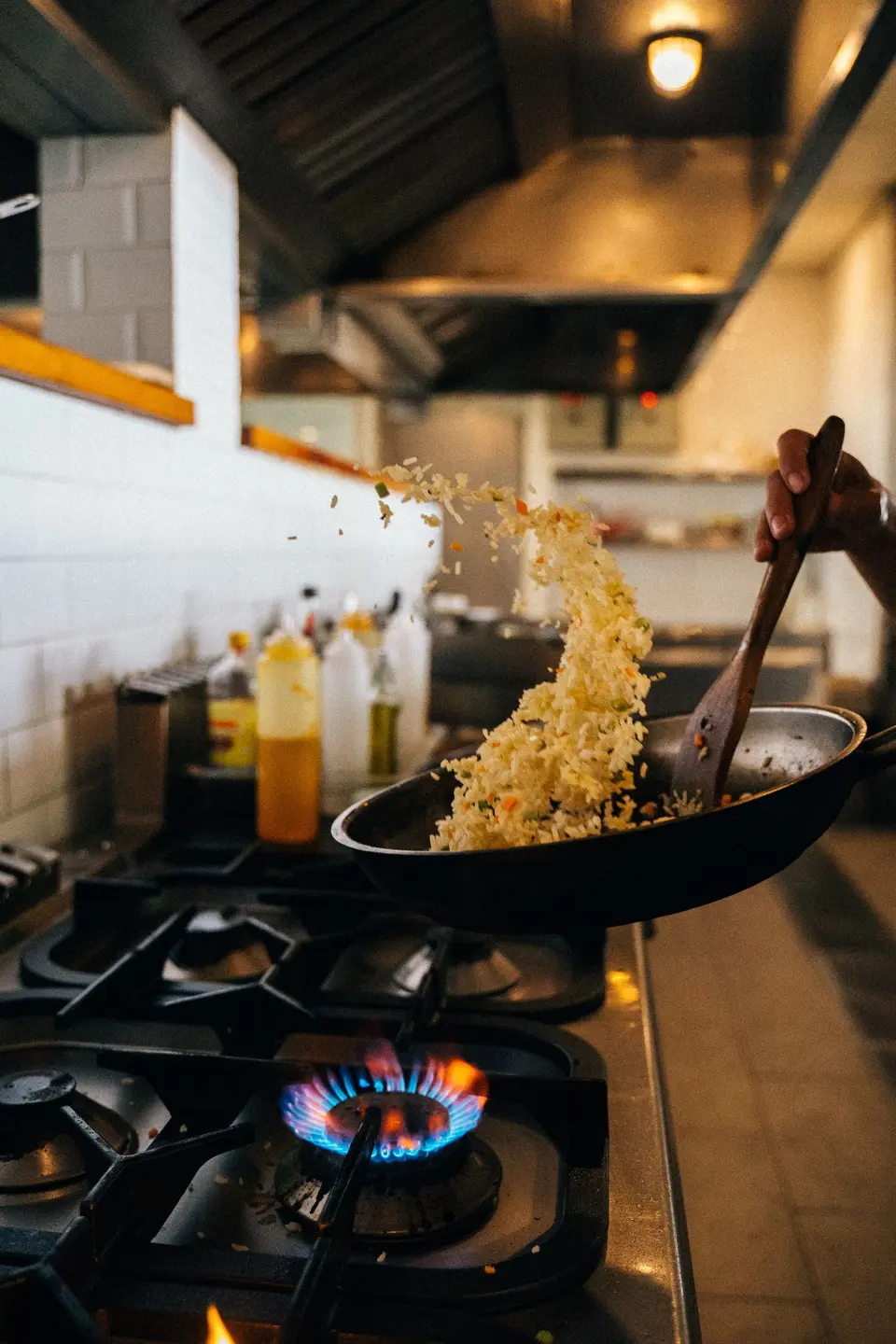 Rice tossed high out of a wok over a gas flame in a white-tiled kitchen